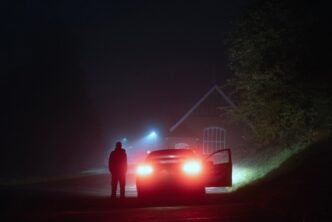 A mysterious person standing next to a car on a spooky empty road on a foggy night. Silhouetted by street lights.