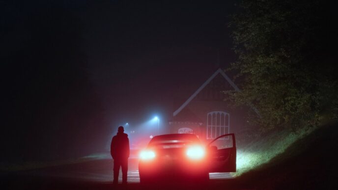 A mysterious person standing next to a car on a spooky empty road on a foggy night. Silhouetted by street lights.